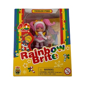 May include: A Rainbow Brite Tickled Pink doll in a yellow box with rainbow borders. The packaging features the text 40 Years Of, Rainbow Brite, and a warning label. The doll has pink hair, a white fur-trimmed hood, and a colorful outfit.
