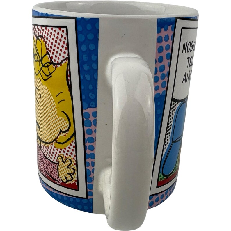 May include: White ceramic mug with a comic book style design. The mug features a yellow cartoon character on one side and the text "NOBODY TELLS ANY" on the other. The mug has a white handle and a blue rim with pink polka dots.