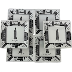 May include: Six square white ceramic plates with black New York City skyline designs. Each plate features a central square with a skyscraper and the words "New York". The borders showcase iconic landmarks.