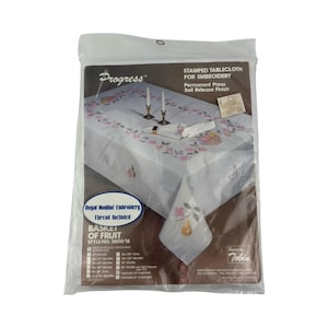 May include: A packaged tablecloth for embroidery, featuring a "Basket of Fruit" design. The white cloth has a floral border and a central fruit basket motif. The package includes thread and instructions for embroidery.