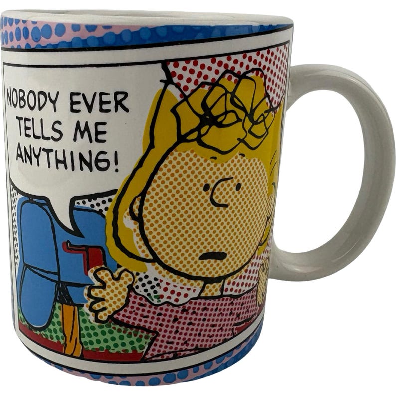 May include: White ceramic mug with a comic book design. It features a cartoon character and the text "NOBODY EVER TELLS ME ANYTHING!". The mug has a white handle and a blue, pink, and red patterned rim.