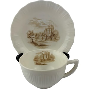 May include: Cream-colored teacup and saucer set. The cup and saucer feature a brown transferware design of a castle scene. The saucer has a scalloped edge. The cup has a handle and vertical ribbed detailing.