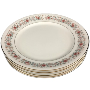 May include: A stack of four white ceramic plates with a floral border design. The border features pink and green flowers with gold trim. The plates are stacked on top of each other.
