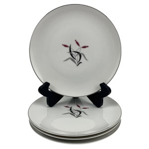 May include: A stack of white ceramic plates featuring a central graphic design of stylized black leaves and three small maroon cattail-like buds. The top plate is displayed upright on a black plastic stand, showcasing the botanical motif clearly.