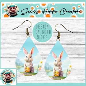 May include: A pair of teardrop-shaped earrings featuring a light blue background with a white rabbit holding a yellow patterned egg. The earrings have gold-toned metal hooks and include the text DESIGN ON BOTH SIDES printed in a circular graphic.