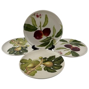 May include: Four white ceramic plates with fruit and leaf designs. The plates feature illustrations of figs, peaches, and leaves in shades of green, red, and brown. The plates are arranged on a white background.