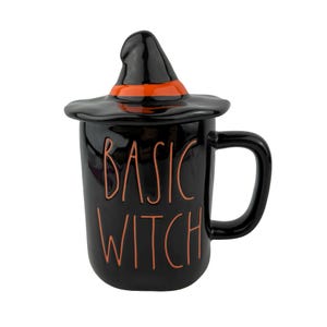 May include: A black ceramic mug with a matching lid shaped like a witch hat. The hat features an orange band. The mug has a handle on the right side and the text BASIC WITCH is written in orange lettering on the front of the black glossy surface.