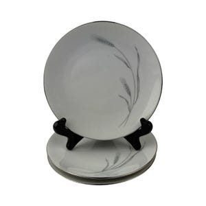 May include: A set of three round white porcelain plates featuring a delicate grey wheat stalk pattern and a thin silver rim. The plates are stacked, with the top plate displayed upright on a black stand to showcase the botanical design.