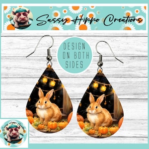 May include: Teardrop-shaped earrings featuring a graphic of a brown rabbit sitting among orange pumpkins with string lights in the background. The design is printed on both sides of the earrings, which hang from silver-toned metal hooks.