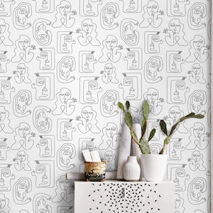 K&ouml;nnte beinhalten: Ein wei&szlig;er Schrank mit zwei T&uuml;ren und einem schwarzen geometrischen Muster. Der Schrank steht vor einer wei&szlig;en Wand mit einem schwarz-wei&szlig;en Linienkunst-Muster von Gesichtern. Ein Hocker steht vor dem Schrank.