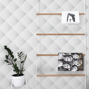 Pode incluir: Um porta-revistas de metal branco com prateleiras de madeira, contendo revistas e livros. O porta-revistas está em pé sobre um piso de madeira clara em frente a uma parede branca com um padrão geométrico. O texto "Buy on patternsColoray Etsy Shop" é visível no canto inferior esquerdo da imagem.