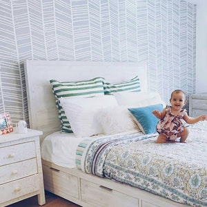 Puede incluir: Una cama blanca con una cabecera blanca y un piecero blanco. La cama tiene un edredón blanco con un patrón floral y una almohada blanca con una almohada azul encima. Una niña pequeña está de pie en la cama con un vestido floral rosa y blanco. La pared detrás de la cama está cubierta con un papel tapiz de patrón de chevron gris y blanco.