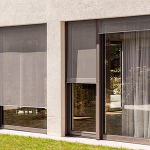 May include: Two large windows with exterior roller shades, one with a sheer curtain behind it. The shades are a light gray color and are partially rolled up. The windows are framed in brown.