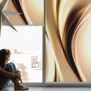 Può includere: Una giovane persona è seduta vicino a una finestra con vista su una città. La finestra è coperta da una tenda a rullo con un motivo marrone e dorato.