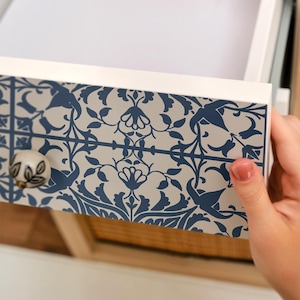 May include: A white drawer front featuring a repeating dark blue floral and vine pattern. The drawer is equipped with a small, round, cream-colored ceramic knob decorated with a dark gray leaf motif on its surface.