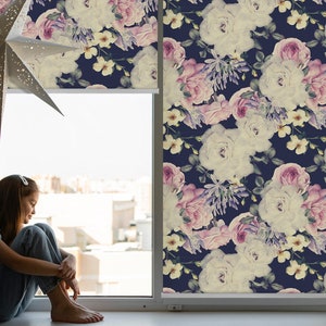 Op de afbeelding: Een raam met een rolgordijn met een bloemenpatroon. Het rolgordijn heeft een herhalend patroon van roze en witte rozen, paarse bloemen en groene bladeren op een donkerblauwe achtergrond. Een jong persoon zit op de vensterbank.