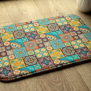 Puede incluir: Una alfombra de baño colorida con un patrón geométrico de cuadrados y círculos en tonos azules, naranjas, amarillos y marrones. La alfombra tiene una superficie texturizada y un borde cosido.