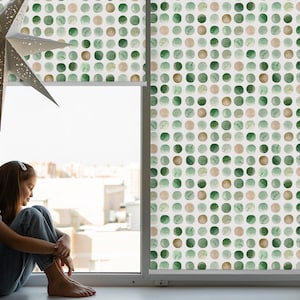 以下が含まれることがあります： 緑と茶色の水玉模様の窓のシェードのある窓のそばに座っている若い人。シェードは窓枠の上まで下げられています。