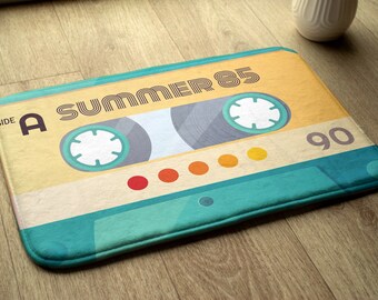 Könnte beinhalten: Eine türkisfarbene und gelbe Badematte mit einem Vintage-Kassettenband-Design. Das Band hat den Text "Side A Summer 85" und "90" aufgedruckt.