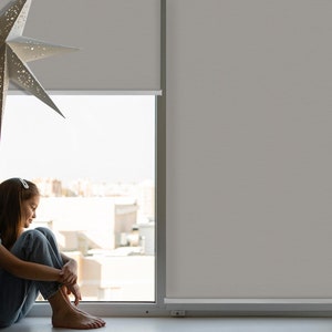 Puede incluir: Una persona joven se sienta junto a una ventana, mirando la ciudad. Lleva pantalones vaqueros y una camisa blanca. Una decoración blanca en forma de estrella cuelga del techo.