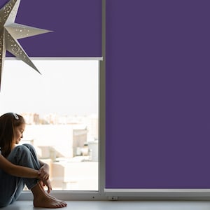 May include: A young person sits by a window with purple blinds, looking out at the city. The person is wearing jeans and a white shirt.