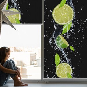 Puede incluir: Una persiana enrollable negra con un diseño de fruta de lima. Las limas caen a través del agua y salpican. La persiana está parcialmente enrollada, revelando una ventana con vista a una ciudad.