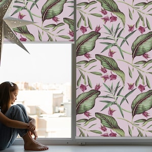 以下が含まれることがあります： ピンクの花柄のブラインドが付いた窓。ブラインドには緑の葉とピンクの花が描かれています。窓際に若い人が座っています。