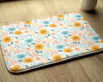 May include: A white bath mat with a floral pattern featuring blue, orange, and pink flowers. The mat has a white background and a light gray border.