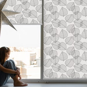 May include: A young person sits by a window with a white background and black leaf pattern roller shade. The window is open and the person is looking out the window.