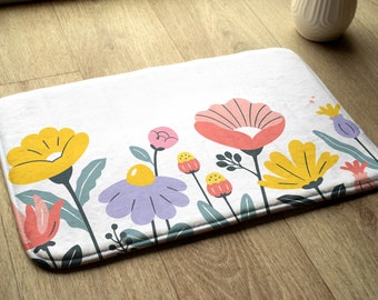 May include: A white bath mat with a colorful floral design. The flowers are yellow, pink, purple, and green. The mat has a white background and a white border.