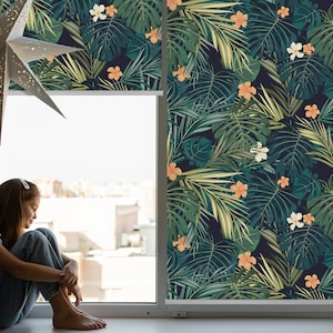 Puede incluir: Una ventana con una persiana enrollable de estampado tropical. La persiana tiene un fondo verde oscuro con grandes hojas verdes y flores naranjas y blancas. Una persona está sentada en el alféizar de la ventana mirando hacia afuera.