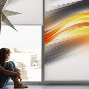 Peut inclure: Une jeune personne est assise près d'une fenêtre avec vue sur des bâtiments au loin. La fenêtre est équipée d'un store enrouleur blanc avec un motif de vagues orange et jaunes sur fond gris.