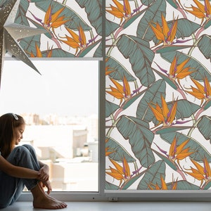May include: A window with a white background and a floral pattern. The pattern features orange and green tropical flowers and leaves. A young person is sitting on the windowsill.