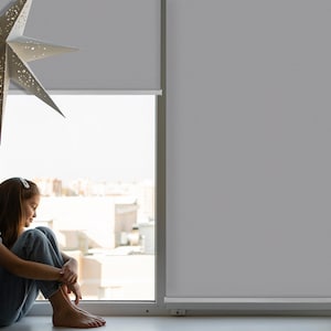 以下が含まれることがあります： 窓際で座って街を見ている若い人。白いシャツと青いジーンズを着用しています。窓は白いブラインドで覆われています。