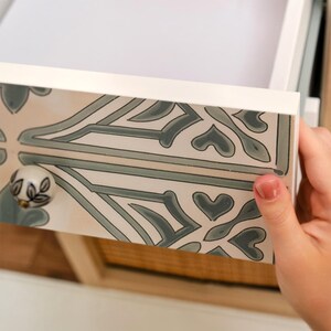 Puede incluir: Cajón blanco con frente decorativo con un patrón repetido en tonos grises y beige. El cajón se está abriendo, revelando el interior. Se ve un pomo decorativo con un diseño floral.