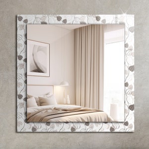 May include: A square mirror with a white frame featuring a brown floral pattern. The mirror reflects a bedroom interior with a bed, curtains, and a window.