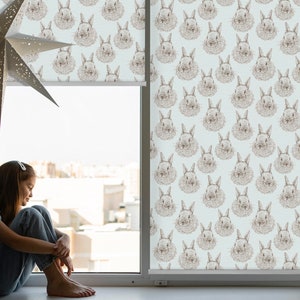 Peut inclure: Une jeune personne est assise près d'une fenêtre avec un store enrouleur blanc orné d'un motif répété de visages de lapins bruns. Le store est en partie enroulé, révélant une vue sur des bâtiments au loin.