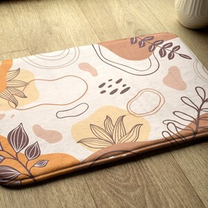 May include: A white bath mat with a brown, orange and beige abstract floral pattern. The mat has a textured surface and a brown border.