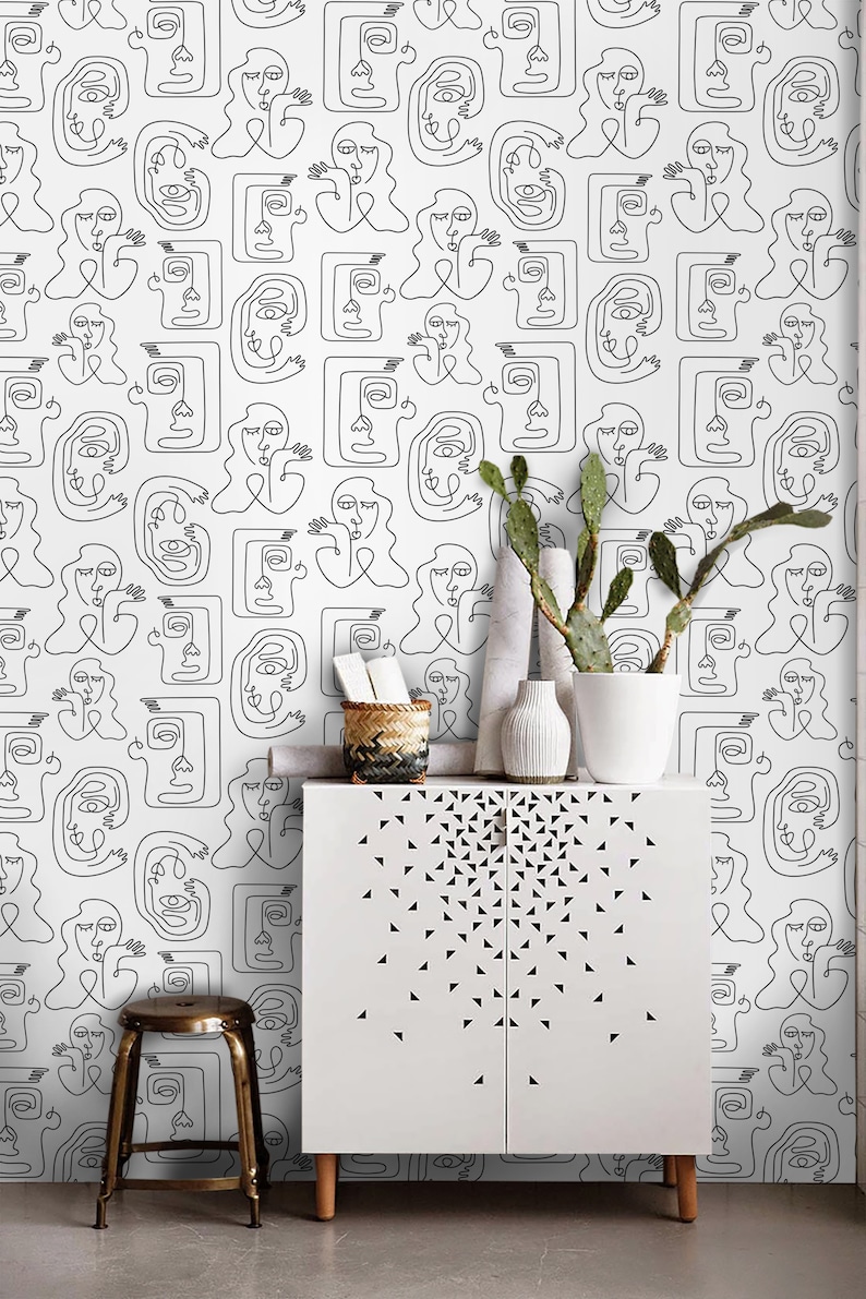K&ouml;nnte beinhalten: Ein wei&szlig;er Schrank mit zwei T&uuml;ren und einem schwarzen geometrischen Muster. Der Schrank steht vor einer wei&szlig;en Wand mit einem schwarz-wei&szlig;en Linienkunst-Muster von Gesichtern. Ein Hocker steht vor dem Schrank.