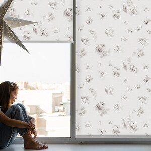 以下が含まれることがあります： 白いローラーシェードがついた窓のそばに若い人が座っています。シェードには、水彩画のような月の模様が繰り返し描かれています。シェードは部分的に巻き上げられており、遠くの建物が見えています。