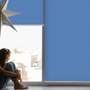 Könnte beinhalten: Eine junge Person sitzt an einem Fenster mit blauen Jalousien und blickt auf die Stadt. Die Person trägt Jeans und ein weißes Hemd.