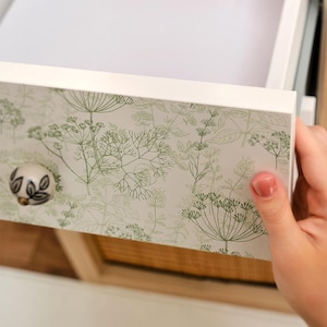 Peut inclure: Un tiroir blanc avec une impression botanique de feuilles et de tiges vertes. Le tiroir est ouvert, révélant l'intérieur. Un bouton décoratif avec un motif floral ajoute une touche d'élégance à l'avant du tiroir.