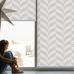 May include: A young person sits by a window with white blinds featuring a geometric pattern. The person is looking down and has their legs crossed. The window is open and there is a view of buildings outside.