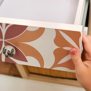 May include: White drawer with a floral pattern in shades of rust, peach, and cream. The drawer pull is a small, round knob with a floral design. The drawer is being opened.