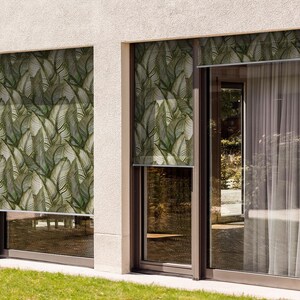 May include: Two windows with white blinds featuring a green and white tropical leaf pattern. The windows are on a white wall with a light brown trim.