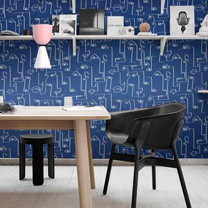 May include: A white table with a black stool and a black chair sits in front of a wall with a blue and white abstract pattern. The pattern features white lines forming faces and eyes on a blue background. The wall has white shelves with books and decorative objects.