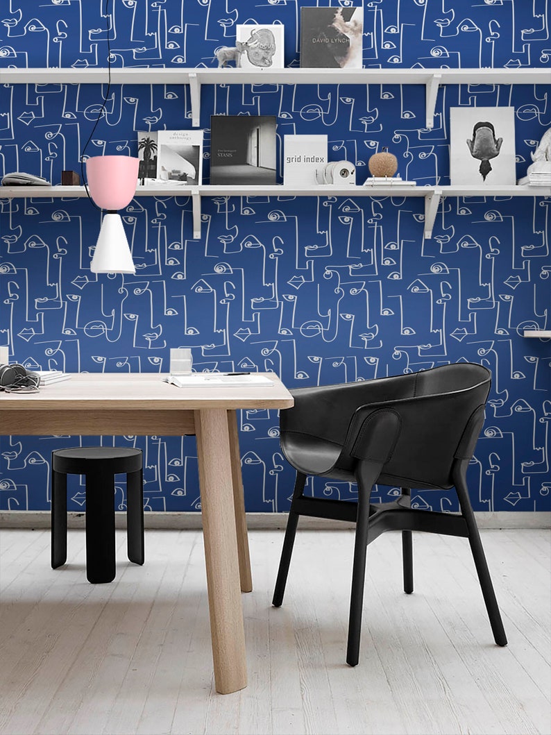 May include: A white table with a black stool and a black chair sits in front of a wall with a blue and white abstract pattern. The pattern features white lines forming faces and eyes on a blue background. The wall has white shelves with books and decorative objects.