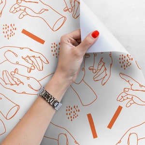 Pu&ograve; includere: Carta da regalo bianca con un motivo ripetitivo di disegni di mani arancioni e forme astratte. Una mano sta staccando la carta per rivelare pi&ugrave; del motivo.