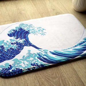 Könnte beinhalten: Eine weiße Badematte mit einem blauen und türkisfarbenen Wellendesign. Das Wellendesign erinnert an die Große Welle vor Kanagawa, einen berühmten japanischen Holzschnitt.