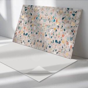 May include: A rectangular decorative panel with a terrazzo design. The panel features a light beige background with colorful flecks of blue, yellow, orange, and green. A white sheet of paper is in front of the panel.